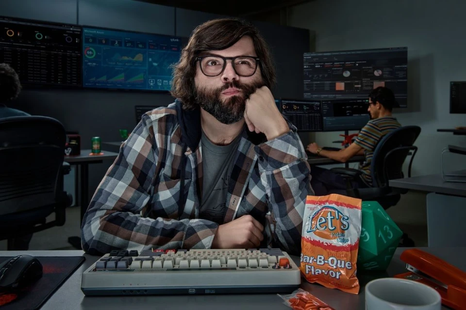 Man with glasses, long hair, and beard leans at a desk with a keyboard, chips, and a multi-monitor workstation in the background.